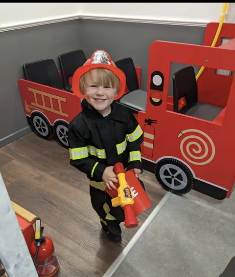 A child dressed as a firefighter beside the little red fire engine at Let’s Pretend
